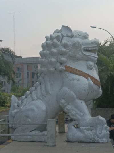 A static, eye-level image captures a large, white stone statue of a Chinese guardian lion, or 'foo dog,' prominently situated on a low, rectangular base. The statue, depicting a male lion, faces right with its mouth agape, revealing stylized teeth and gums. Its body is adorned with a textured, segmented mane and a patterned band resembling a belt around its midsection. The front right paw of the lion rests upon a small, spherical object or cub. The statue exhibits signs of weathering, including surface discoloration and minor erosion.
In the immediate foreground, a two-bar metal railing runs horizontally, obscuring the lower portion of the statue's base. To the far right, partially obscured by the statue's lower section, a single individual wearing dark clothing is seated or crouching on the ground. Their face and specific actions are not discernible.
The background features lush green foliage, including palm trees, on both the left and right sides of the statue. Behind the vegetation on the left, a multi-story building with a light-colored facade and visible windows is present. Further in the background, a taller, dark antenna structure rises against a hazy, light grey sky. To the upper right, a modern streetlamp with a curved arm extends over the statue.
This scene is located outside Vihara Dharma Bhakti temple in Glodok, West Jakarta, within the Special Capital Region of Jakarta, Indonesia.