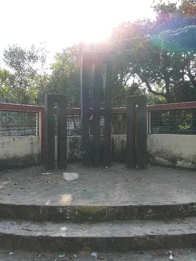 A concrete monument stands in Shibpur, Bangladesh, under strong sunlight, casting a lens flare from the upper center. The monument consists of five dark, vertical rectangular pillars of varying heights, arranged on a raised, circular concrete platform. The leftmost pillar and a partially visible rightmost pillar each feature a rectangular cutout near their tops. Two concentric concrete steps lead up to the platform. The ground, steps, and platform are visibly dirty with scattered debris, including dried leaves, small paper fragments, and a crumpled white plastic object. Behind the monument, a low concrete wall with dark staining is visible, topped by a red-painted concrete beam and a metal grid fence. Dense green foliage and trees form the background, illuminated by the bright sun. No individuals are present.