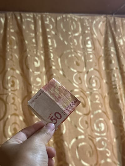 A close-up, eye-level, vertical shot captures a hand holding a folded banknote. The banknote is a light brown color with reddish-orange and golden-yellow accents. Prominently displayed on the visible portion of the banknote are the large red numerals "50" and text in a script that appears to be Cyrillic. To the left of the "50," the words "КЫРГЫЗ БАНКЫ" are visible. To the right of the "50" and slightly above, the Cyrillic letters "ЭЛУУ СОМ" are visible. The background consists of a tan, textured curtain with a swirling, embossed pattern. The lighting suggests an indoor setting. No specific location in the city of International, Kyrgyzstan is depicted, only the banknote and the background curtain.