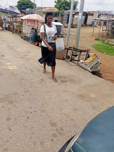 A woman walks on a paved road, with a black skirt and a white t-shirt. She carries a black bag on her shoulder and a white plastic container in her right hand. Behind her, stalls line the side of the road, some covered by umbrellas. One stall has a sign that says "PUS AVAILABLE". In the background, there are tin shacks and greenery. A man sits at one of the stalls. The scene appears to be a market or roadside settlement. The sky is overcast, suggesting it could be daytime, possibly after rain or before a storm. The overall impression is one of everyday life in a busy, possibly developing, area.
