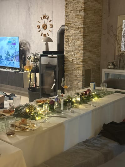 A living room scene includes a stone-clad fireplace with a modern glass-fronted stove. A clock hangs on the wall next to the fireplace. A television displays the text "ИКСЫ! 2025." A long dining table is set with festive decorations, including greenery, lights, glasses, and plates with leftover food. The table extends towards a cushioned seating area or sofa.