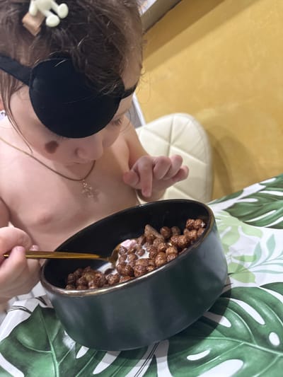 A child wearing a black sleep mask and a gold necklace is holding a bowl of cereal with milk and a gold spoon. The bowl is dark green with a gold rim. The background includes a yellow wall, a white chair, and a green and white patterned curtain. The location is unspecified.