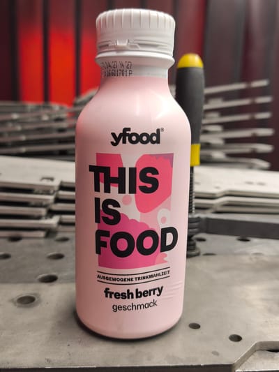 A close-up, eye-level shot captures a pale pink bottle of "yfood THIS IS FOOD" brand beverage, positioned on a grey, metallic surface. The bottle has a white cap with a textured grip and bears black text with pink graphic elements. Visible text on the bottle includes "yfood", "THIS IS FOOD", "AUSGEWOGENE TRINKMAHLZEIT", "fresh berry", and "geschmack".

Behind the bottle, a black tool with a yellow cap is partially visible, positioned upright. The background features blurred, red vertical stripes on the left and rows of stacked, grey metal objects, possibly plates or sheets, on both sides and in the background, suggesting an industrial or workshop environment. The lighting is even, highlighting the product. The image does not depict any specific identifiable landmarks or features definitively placing it within the city of Bad Honnef, Germany, beyond the general nature of its contents and potential for it to be a product sold there. The scene is static and shows no human presence or activity.