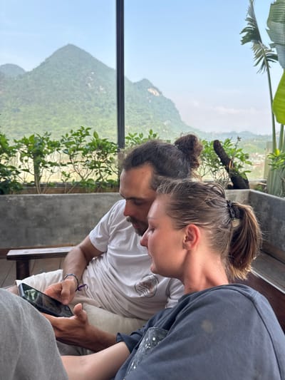 The image, oriented correctly, depicts two individuals intently focused on a smartphone in a scenic indoor setting. In the foreground, a female with her hair tied in a ponytail wears a dark long-sleeved top and holds a black-cased smartphone, her gaze directed downwards at the screen. Behind her and slightly to the left, a male with dark hair in a bun and a beard also leans in, looking at the phone. He wears a white t-shirt with a faint, indecipherable logo and a purple bracelet on his left wrist.
The two appear to be sharing or viewing content on the device. They are seated, likely on a bench, next to a modern concrete ledge or wall that separates them from a stunning panoramic view. Outside, through large windows or an open balcony, lush green mountains dominate the landscape. The sky is bright and clear, suggesting daytime, with a soft haze over the distant peaks. Green foliage, including large palm-like leaves, is visible close to the viewing area and further out on the mountain slopes, indicating a verdant environment typical of Hà Giang 1 Ward, Vietnam. The scene conveys a moment of shared concentration amidst a beautiful natural backdrop. No specific text is clearly legible in the image.