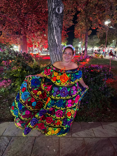 A smiling woman in a colorful, off-the-shoulder dress with intricate floral embroidery stands outside at night. She wears a floral headband, and the dress has multiple tiered layers. Behind her are trees with reddish foliage, greenery, and street lights. The background is blurry, suggesting a public space. To her right, a paved area with stone tiles can be seen.