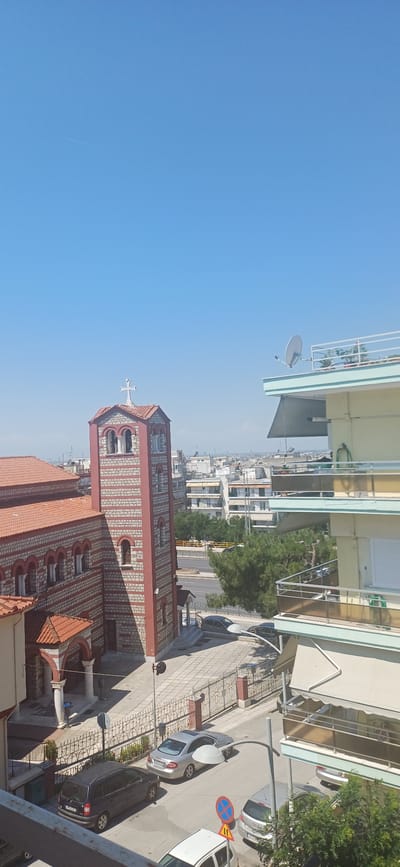 This is a medium-angle, eye-level shot of a street scene in Stavroupoli Municipal Unit, Greece, on a clear, sunny day. The foreground features a road with parked cars, including a dark minivan on the left and a silver sedan to its right. A white vehicle is partially visible at the bottom. Two street lamps are prominent, with one arching over the road. A red and white striped church with a red tile roof and a bell tower topped with a white cross is on the left. Adjacent to the church is a multi-story building with balconies and a satellite dish on the roof. The background shows more urban architecture, trees, and a clear blue sky with a faint contrail from an airplane.