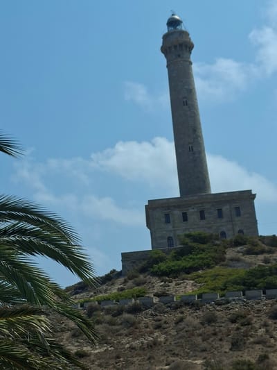 The image shows the Cabo de Palos Lighthouse, a tall, cylindrical lighthouse made of stone, situated on a hill. The lighthouse has a rectangular base with several windows. In the foreground, palm fronds are visible, obscuring part of the view. The background consists of a clear blue sky with scattered clouds and the dry, shrub-covered hillside leading up to the lighthouse. The scene is an outdoor, daytime shot of a coastal landmark.