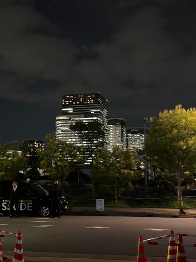 In the city of Koto, Japan, at nighttime, a street scene unfolds with a dark sky illuminated by city lights. The foreground features a black taxi with illuminated signage reading "SIDE" and Japanese characters. Two individuals, appearing to be men in dark attire, stand near the taxi; one to the left appears to be a passenger about to enter, while the other on the right stands by the open rear passenger door. Traffic cones and barriers line the road. Behind them, a row of trees with glowing green foliage separates the street from a backdrop of modern, illuminated high-rise buildings. The buildings display numerous lit windows, suggesting occupancy and urban activity. The overall atmosphere is one of urban nightlife.