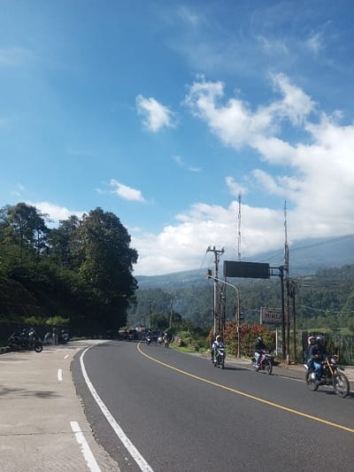 The image displays a two-lane asphalt road winding through a mountainous and heavily vegetated landscape in Ka Thaung (Upper), Myanmar, under a bright blue sky with scattered cumulus clouds. The road features a solid yellow center line and white edge markings. On the left side of the road, a concrete pull-off area accommodates approximately 5-7 individuals standing near several parked motorcycles. A steep, densely tree-covered hillside rises immediately behind this area. On the right side of the road, multiple motorcycles are in motion, heading away from the viewer. In the immediate foreground, a dirt bike carries two helmeted riders, the lead rider in a green helmet and jacket. Behind them, additional motorcyclists are visible, all wearing helmets. The right shoulder of the road contains utility poles with overhead lines, two tall communication towers, and a partially obscured white sign. The background reveals multiple layers of green, forested mountains, with a faint wisp of smoke visible rising from a distant valley on the right.