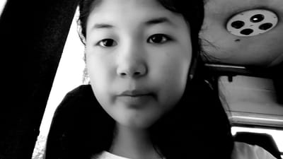 A black and white close-up selfie captures a young person's face, positioned slightly off-center. The individual's gaze is directed towards the camera, with their eyebrows slightly raised. Their lips are closed, and their nose is centrally located on their face. The background is dark, with a bright light source visible on the upper right side of the image. A circular object with four dark circles is mounted on what appears to be a ceiling. The person's hair is dark and styled in pigtails that frame their face. The lighting creates harsh shadows, particularly on the left side of the face. The image is grainy, indicating a low-resolution capture or low light conditions. The overall impression is of an informal snapshot taken from a personal device. There is no indication of Kaarman, Kyrgyzstan.
