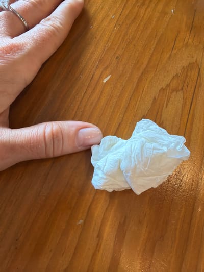 A close-up, overhead view depicts a light-skinned human hand and a crumpled white tissue resting on a wooden surface. The hand enters the frame from the top-left, extending downwards. The thumb, displaying a light-colored, manicured nail, is positioned near the center-left. A silver-colored ring with a single clear stone is visible on a finger (likely the ring finger) partially cropped in the upper-left quadrant. To the immediate right of the thumb, a single crumpled white paper tissue with visible creases is situated on the surface. The underlying environment is a light brown wooden surface, likely a table, displaying distinct wood grain patterns running diagonally and several small, light-colored debris particles. The specific location within Benoa, Indonesia, cannot be determined from the visual information provided in the image.