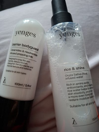 Depicted are two cosmetic product bottles resting on a light-colored, folded textile surface. The bottle on the left is an opaque white, cylindrical container with a black base, labeled "yenges" and identified as "barrier bodyguard," a "ceramides & niacinamide infused prepping milk," with a stated volume of "100ml / 3,4oz." The bottle on the right is a clear, cylindrical container with a black spray cap, also branded "yenges" and named "rice & shine," containing a bubbly, translucent liquid described as "Oryza Sativa (Rice) infused water," with a stated volume of "150ml." The background consists of soft-focus, light-toned fabric. No individuals are present. The specific geographic location in the city of Selyanin, Bulgaria, cannot be determined from the visual evidence presented in the image.