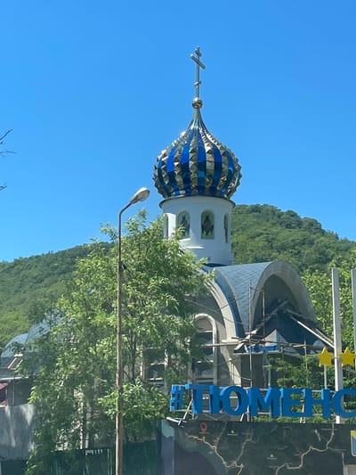 The image captures a clear, bright day with a vibrant blue sky dominating the upper portion. In the foreground, a street lamp stands tall, its white head angled downwards. Partially obscured by lush green trees and foliage, the lamp post is weathered and shows signs of age.
Behind the trees, the domed roof of a building, likely an Orthodox church, is visible. The dome is a striking feature, adorned with alternating panels of blue and gold, culminating in a cross at its apex. The church itself appears to be under some form of construction or renovation, as scaffolding is visible around its structure, particularly near the blue-tiled roof. The building's facade features white architectural elements and arched windows.
Further back, a lush, green hillside covered in dense trees rises behind the church. The overall environment suggests a semi-urban or suburban setting where nature is in close proximity to man-made structures.
In the lower right portion of the image, large blue letters form part of a sign that reads "ТЮМЕНСКОЙ", which translates to "Tyumenskiy" in Russian. This directly indicates the location as being within the Tyumenskiy region of Russia. Below this sign, a dark surface displays a map-like design with various markings and symbols, and a Ukrainian flag is visible on the far right.