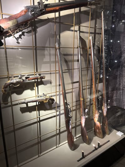 The image displays a collection of antique firearms presented in a museum exhibition. The firearms are mounted on a light-colored wall with a grid of thin metal rods.
In the foreground, there are two ornate pistols displayed horizontally, one above the other. Both pistols feature intricate metalwork on their stocks and barrels, with wood and metal components. The upper pistol is slightly larger and more elaborately decorated.
Behind the pistols, several long guns are displayed vertically. These appear to be rifles or muskets, also with wooden stocks and metal barrels and mechanisms. Some of these long guns have visible ornate decoration on their stocks and metal parts.
The background wall is a neutral, light gray color, with shadows cast by the firearms and the display structure. A glass display case likely encloses the artifacts. Reflections on the glass indicate the presence of lighting and possibly other exhibits in the museum.
A sign with Cyrillic text is partially visible on the right side of the image, suggesting the exhibition is in Russia. Given the context of antique firearms and Russian text, this collection could be part of the Gatchina Palace Museum in Gatchina, Russia, which houses significant historical armaments.