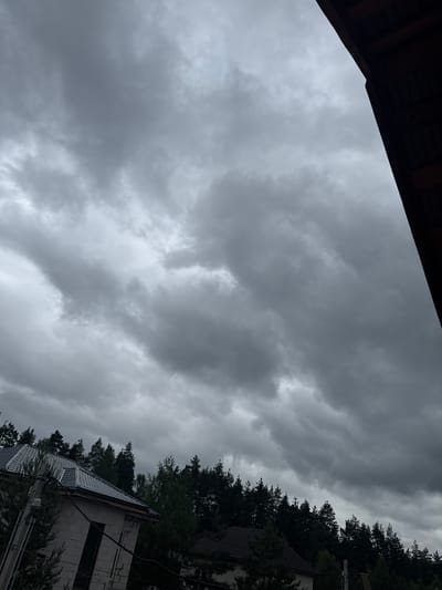 This is a vertical, low-angle shot of a cloudy sky over a forested area and a partially constructed building, taken in Protasovo, Russia. The sky is filled with heavy, dark grey clouds, suggesting overcast or stormy weather. The time of day is not clearly discernible due to the cloud cover, but it appears to be daytime.
In the foreground, a dark, wooden structure, possibly an overhang or roof, occupies the upper left corner. The middle and right portions of the image are dominated by the dense, dark green canopy of coniferous trees, stretching along a diagonal line from the mid-right to the top right.
Below the trees, a portion of a building is visible. It appears to be under construction, with grey concrete blocks forming its walls and a dark grey, patterned roof. The roof has a modern, tiled appearance. Power lines and utility poles are also visible in front of the building and trees, indicating a semi-rural or suburban setting.
There are no people visible in the image, and no text is present. The overall mood is somber and perhaps a little dramatic, due to the dark clouds and the unfinished state of the building.