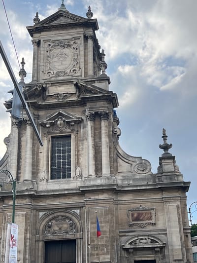 A low-angle shot captures the facade of Saint-Servais Church (Sint-Servatiuskerk) in Saint-Josse-ten-Noode, Belgium. The stone building, rendered in light grey, features intricate classical architectural elements. The central portion of the facade is dominated by a large, ornate window with a grid pattern, framed by columns and topped with decorative stonework. Above this, a pediment contains a carved crest, and higher still, a circular relief depicting a cherub and a radiant heart is set within an elaborate frame. The upper part of the facade culminates in a stepped roofline adorned with finials and decorative urns. To the left, a street lamp with a curved arm is visible, along with a vertical banner displaying red and white text. A small Belgian flag, red, yellow, and black, flies from a pole on the right side of the entrance. The sky above is partly cloudy, with patches of blue visible through white and grey clouds.