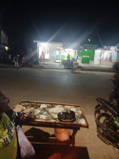 This nighttime image depicts a commercial street in Kihara ward, Kenya. In the immediate foreground to the left, a person with dark skin is partially visible, wearing a light-colored top and a patterned bag, with an open-mouthed expression. Adjacent to this individual is a rectangular wooden table with a light-colored, possibly tiled, surface. On the table, a dark bowl and multiple metallic implements are present. A reddish glow beneath the table indicates a heat source, and a white bucket is situated underneath. To the right, the rear wheel and exhaust of a motorcycle, featuring a green side panel, are visible.

The midground consists of a paved or dirt road. Several indistinct figures are distributed across the scene; one individual is walking on the left side of the road, while others are congregated near the shops in the background. At least one additional motorcycle is parked on the left side of the road.

The background features a brightly lit row of single-story commercial buildings. From left to right, a shop displaying hanging items, a produce stall with patrons, and a Safaricom/M-PESA agent location characterized by prominent green signage are discernible. Another storefront is partially visible on the far right. Steps lead up to these business entrances. Multiple artificial light sources from the establishments create significant lens flares in the upper portion of the image, set against a dark night sky.