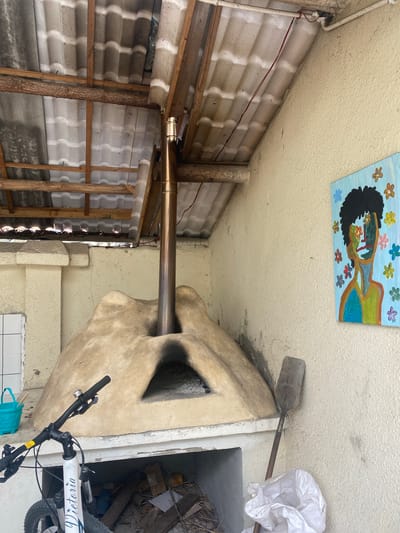 An outdoor covered area features a rustic scene. A domed, clay pizza oven with a stainless steel chimney dominates the center of the frame. To the left, a white bicycle with black handlebars is parked. A teal plastic bucket rests beside a white tiled surface. On the right wall, a vibrant, abstract painting of a face with flowers for eyes hangs. A shovel leans against the wall next to a white plastic bag. The roof is constructed with wooden beams and corrugated panels. Debris is visible in an opening beneath the pizza oven. The location is identified as Dujiangyan City, China.