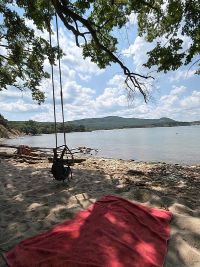 The image depicts a serene beach scene in Dyuni, Bulgaria, on a sunny day. In the foreground, a vibrant red towel is spread on the sandy shore, bathed in dappled sunlight filtering through tree branches. Behind the towel, a backpack hangs from a rustic swing made of thick ropes suspended from a large tree. The swing overlooks a tranquil body of water, likely the Black Sea, with gentle waves lapping against the pebbled shoreline. In the distance, lush green hills form a picturesque backdrop against a sky filled with fluffy white clouds. The overall atmosphere is one of peaceful relaxation and natural beauty.