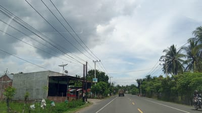 The image depicts a road scene in the vicinity of Blangtambue, Indonesia. A paved road with yellow lane markings stretches into the distance, flanked by lush green vegetation and palm trees. Overhead, a complex network of power lines crisscrosses the sky, supported by utility poles. On the left side of the road, a building with a grey concrete facade and a covered outdoor area is visible. This building has two satellite dishes mounted on its roof. Further down the road, a truck is traveling away from the viewer. On the right side of the road, a person on a motorcycle is also moving away from the camera. The sky is partly cloudy, with a mix of grey and white clouds. The overall impression is that of a rural or semi-rural road in a tropical climate.