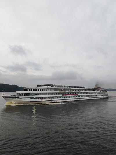 A wide-angle, eye-level, full shot captures a large, white passenger ship with multiple decks sailing on a dark, choppy river under a heavily overcast sky. The ship, identified by the Cyrillic inscription "НИЖНИЙ НОВГОРОД" (Nizhny Novgorod) on its bow and "НИЖНИЙ НОВГОРОД" along its side, has a red stripe running along its hull and a red stripe along the top deck. A plume of black smoke billows from a stack at the rear of the ship. The ship is creating a wake of white foam at its bow and stern. The background consists of a dark, tree-lined shoreline on the left and a distant, hazy shoreline on the right. The water reflects the ship's lights and the overcast sky. The overall atmosphere is somber and muted. The image is not specifically located in Khoroshevo, Russia; the text on the ship refers to the city of Nizhny Novgorod.