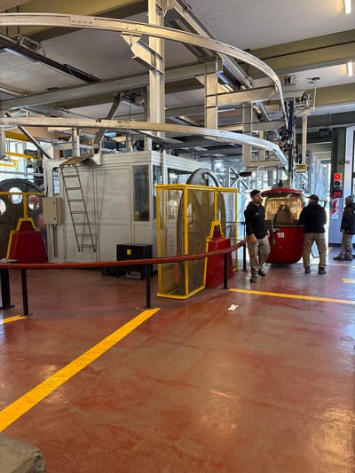 The image displays the interior of a cable car station, identified as being in San Carlos de Bariloche, Argentina. The floor is reddish-brown, marked by yellow linear demarcation strips and scuff marks. A small piece of white debris rests on the floor in the mid-ground. Overhead, a robust industrial structure of interconnected grey metal beams, tracks, and mechanical components supports the cable car system.

A red gondola, numbered "18," is positioned at a boarding point in the mid-ground. Its windows are tinted, revealing indistinct reflections of occupants or interior structure. Three adult males, consistent in appearance with station operators, are present. The male closest to the left is facing right, standing near a yellow metal mesh safety barrier protecting a large industrial wheel. Another male stands with his back partially to the camera, positioned at the open entrance of the gondola. A third male stands further right, facing left towards the gondola, next to a wall-mounted fire extinguisher and a red digital display indicating "05."

To the left and rear of the gondola, a light grey, rectangular control or utility structure stands, against which a metal ladder is leaned. An electrical box is mounted on its exterior. A black box with a yellow warning sticker is on the floor in front of this structure. An orange safety rope barrier, supported by black stanchions, spans the foreground. Natural light enters from a large opening or window on the far right side of the scene.