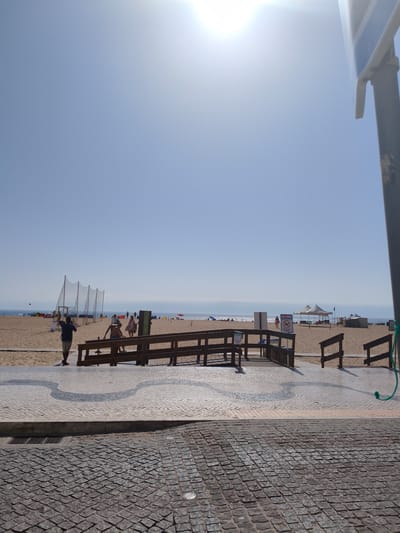 The image shows a beach scene in Nazaré, Portugal, on a sunny day. In the foreground, there is a cobblestone walkway with a decorative wavy pattern. A wooden ramp with railings leads up to the beach. On the beach, several people are visible, some walking and others relaxing under umbrellas. There are also sports nets set up on the sand, possibly for volleyball or a similar activity. In the background, the ocean stretches out to the horizon under a clear blue sky. The sun is bright, casting a glare in the upper right portion of the image. There are also some white tents set up on the beach.