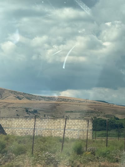 This media file captures a vertical, wide-angle view of a landscape and sky. The left side of the image is dominated by a cloudy sky with a bright sunlit area. Two faint, white contrails are visible in the sky, suggesting the presence of aircraft. The right side of the image shows a terrestrial landscape, divided into a dry, grassy hillside and a strip of green vegetation.

The hillside is a light brown or tan color, with sparse vegetation and some darker patches that might be shrubs or shadows. It appears to be a sloping terrain, possibly a rural or natural setting. Adjacent to the hillside is a paved area, made of light-colored rectangular stones arranged in a brick-like pattern. This paved area is bordered by a series of vertical, dark poles, spaced at intervals. Beyond the paved area and poles, there is a strip of green grass with scattered white wildflowers.

In the distance, along the top edge of the hillside, a dark line of trees or dense vegetation can be seen. There are also some utility poles visible on the hillside in the far distance, suggesting infrastructure in the area. The lighting indicates it is daytime, with bright sunlight, though the exact time is not specified. The presence of clouds suggests a partly cloudy sky. The overall scene conveys a sense of openness and natural beauty, possibly a rural or undeveloped area. There is no visible text in the image.

The image appears to be taken from a moving vehicle, possibly a car, given the slight blur and the angle of the shot which seems to be looking out a window. The reflection of what appears to be a car's interior is faintly visible on the left side of the image, particularly in the upper left quadrant.