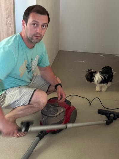 The image shows a man sitting on the floor next to a red and black vacuum cleaner. He is wearing a light blue t-shirt with a graphic design and light grey shorts. The man has short, dark hair and stubble. He is looking towards the camera with a surprised expression. To the left of the man, a small, fluffy black and white dog is sitting. The scene appears to be indoors, possibly in a room under renovation or being cleaned, as evidenced by the unfinished wall on the left and the concrete floor. A dark electrical cord from the vacuum cleaner snakes across the floor. The lighting suggests it is daytime. There is no visible text in the image.