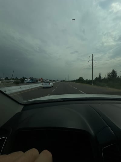 This media file captures a view from inside a vehicle traveling along a highway in Sergeli, Uzbekistan. The scene depicts a cloudy, overcast sky, suggesting potentially inclement weather or a late afternoon/early morning time of day. The road ahead is visible, with a white vehicle in the distance and construction barriers lining the right side of the highway. Power lines and pylons are a prominent feature in the landscape, indicating the presence of infrastructure. The interior of the car is visible on the right side of the frame, with a dark dashboard and a hand in the lower right corner. The overall impression is one of transit through a somewhat industrial or developing area, with a somber sky adding to the mood. No discernible text is visible in the image.