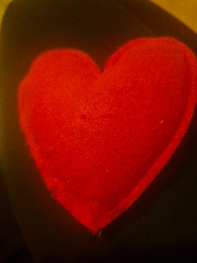 The image displays a single, red, heart-shaped object centrally positioned against a predominantly dark background. The object exhibits a slightly textured, soft appearance, consistent with plush or felt material, and is uniformly vibrant red. The background is a deep, undifferentiated dark tone, possibly black or very dark green/brown, with subtle, blurred hints of a lighter, yellowish or olive hue visible in the upper-left and upper-right corners of the frame. The overall lighting is dim, contributing to a visible grain or noise throughout the image. No individuals, actions, or specific contextual elements are present within the scene. The precise location within the city of Hereford, United Kingdom, cannot be determined from the visual evidence provided.