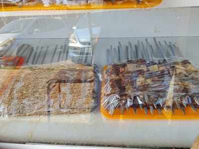 The media file shows a display of food items, likely kebabs, covered in plastic wrap. There are two distinct trays of food, each containing meat skewered on metal rods. The tray on the left contains what appears to be a dry, granular food item, possibly a side dish or a different type of kebab, with a reddish-brown hue and a pebbly texture. The tray on the right holds several skewers of meat, dark brown in color and appearing to be grilled. The skewers are arranged vertically and point upwards, with sharp metal tips visible. Both trays are placed on an orange surface.

Behind the food, the scene is reflective, showing a blurry image of cars. One car, silver in color, is visible on the left side, and another, possibly white or light-colored, is on the right. A license plate with the inscription "AQA" is partially visible on the front of the car on the right. The background appears to be an outdoor setting, with hints of sunlight suggesting it is daytime. The overall impression is that of a food stall or a market display, possibly in Ala-Buka, Kyrgyzstan, given the location context. The reflection on the plastic wrap indicates a somewhat bright environment. No people are visible in the frame.
