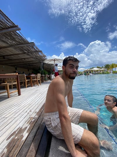 The image shows a man sitting on the edge of a pool in Marina Maroma, Mexico. He has dark hair and a beard and is wearing striped shorts. His torso is bare. Another person is in the pool behind him. The area beside the pool features a wooden deck with seating and a thatched roof structure. The sky is blue with white clouds.