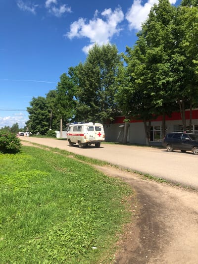 The image depicts a sun-drenched, rural street scene in Izdeshkovo, Russia, on a clear day with scattered white clouds in a bright blue sky. The road is paved and curves to the right, bordered by lush green grass and trees. On the right side of the road, a modern building with a grey corrugated metal facade and red trim stands, partially obscured by several tall, leafy trees. A dark-colored car is parked on the right side of the road in front of the building. On the left side of the road, a white ambulance, bearing a red cross and Cyrillic lettering that appears to read "МЕДИЦИНСКАЯ ПОМОЩЬ" (Medical Assistance), is driving away from the viewer. A white fence runs along the left side of the road behind the ambulance, and further down the road, several people can be seen walking. A dense green bush with pink flowers is visible on the far left of the image. The overall impression is one of a quiet, ordinary day in a small Russian town.