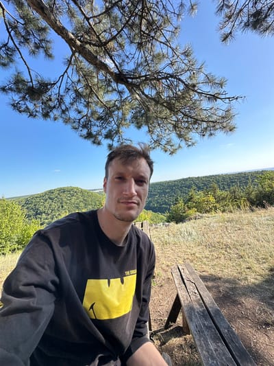A man is taking a selfie outdoors on a sunny day. He is sitting next to a wooden bench. Behind him, there is a lush green landscape with rolling hills and a clear blue sky. A tree branch with pine needles is visible in the foreground, partially obscuring the sky. The man is wearing a black sweatshirt with a yellow graphic print. The location is the city of Nihyn, Ukraine.