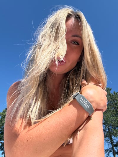 The image is a low-angle, close-up shot of a woman with long, blonde hair against a bright blue sky. Her hair is partially covering her face, obscuring her nose and mouth, but her left eye is visible, looking towards the camera. She is wearing a grey wristband with the text "MOREA RESORT & SPA HOTEL" printed on it, and a small gold charm hangs from it. Her right arm is bent, and her hand is resting on her arm. The background shows the tops of green trees and the clear blue sky. The lighting suggests it is daytime and sunny. The image is an outdoor portrait taken in the city of Anapa, Russia.
