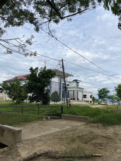 The image depicts a building in Bundar, Indonesia, under a cloudy sky. The building is modern in design, with white walls, blue accents, and a red tiled roof. It appears to be a commercial or institutional building, possibly an office or a small factory, with a parking area in front. Several vehicles, including motorcycles, a yellow car, a white truck, and a tow truck with a crane, are parked in the vicinity. The surrounding area is covered in green grass and trees, with a dirt path leading towards the building. There is a metal fence in the foreground, and a concrete barrier. The overall scene suggests a functional, developed area within the city.