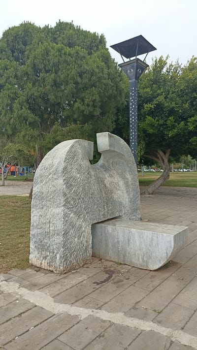 This media file captures an outdoor scene featuring a contemporary stone sculpture in a park-like setting. The sculpture is the primary subject, characterized by its abstract, geometric form with rough-hewn textures and smooth, polished surfaces. It appears to be made of light-colored marble or granite. A large, rounded section forms the base, with a rectangular element extending from it, suggesting a bench or seating area.

Behind the sculpture, lush green trees dominate the background, creating a natural, verdant environment. The foliage ranges from dense, feathery pine-like trees on the left to broad-leafed trees on the right. A distinct lamppost with a dark, pyramidal top and a decorative, patterned base stands to the right of the sculpture, adding an element of urban infrastructure to the natural scenery. The ground in the foreground is paved with rectangular stone tiles, laid out in a pattern. A strip of grass is visible to the left of the paved area, and further in the background, children's playground equipment can be seen, hinting at a public recreational space.

The time of day appears to be overcast or late afternoon, as the lighting is diffused and there are no strong shadows, suggesting a cloudy sky. The overall atmosphere is calm and peaceful. There are no people visible in the frame. There is no discernible text within the image. The location context provided is Mersin, Turkey.