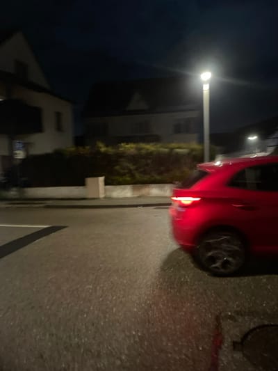 A red car is parked on the side of a dimly lit street at night. A tall, white building with a steeply pitched roof is visible across the street, partially obscured by a green hedge. A street lamp casts a bright glow near the building, illuminating the surroundings. The road is dark gray. A black painted stripe marks the edge of a parking spot. A dark sewer cover is near the car.