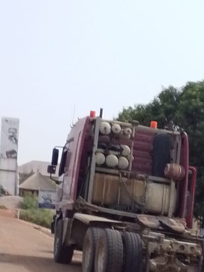 The image shows a heavy-duty truck from its rear-left quarter, captured on what appears to be an unpaved or dirt road in Dungu, Ghana, under bright daylight. The sky is clear, indicating fair weather.

The truck's cab is maroon or reddish-brown, while its chassis and functional components are heavily weathered, displaying metallic gray and yellowish-brown hues. Behind the cab, the truck is extensively fitted with various equipment. Multiple white or off-white cylindrical tanks, possibly gas or air cylinders, are prominently stacked both vertically and horizontally, with at least ten visible. Below these, a larger, yellowish-brown horizontal cylindrical tank is securely fastened. A dark, possibly fabric or rubber, object hangs vertically, and a smaller reddish-brown cylindrical item is positioned on the far right side of the loaded area. Two orange warning lights are mounted atop the structure behind the cab. The truck's rear features at least two sets of dual tires, consistent with a robust heavy-duty vehicle. A rearview mirror is visible on the left side of the cab.

In the background, to the left of the truck, a tall white rectangular billboard or sign is partially visible. The letters "DX" are clearly discernible at the top, accompanied by other smaller, illegible text and a graphic below. Behind this sign, a building with a distinctive conical thatched roof, resembling a traditional African hut or rondavel, is present. Further into the background, lush green trees are visible to the right, and scattered vegetation lines the roadside. A smaller blue sign with a white graphic and some illegible text is also noticeable near the traditional building. No people are visible, and the scene depicts the truck's presence on the road, suggesting a functional, possibly commercial, context.