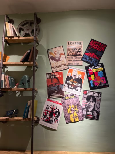 The scene depicts a wall decorated with movie posters, above a rustic-style shelving unit in Xanthi, Greece. The wall is painted a light green. The posters include "Stranger Things," "Easy Rider," "Trainspotting," "Back to the Future Part III," "Chicas Almodóvar," "Once Upon a Time in Hollywood," "Matiador," and "Wednesday Season 2".
The shelving unit is made of dark metal pipes and light wood planks. A film reel is mounted on the wall next to the shelf. Books and a vintage electrical device are placed on the shelves.