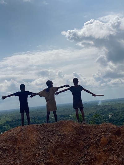 Three young boys stand on a rocky, elevated terrain overlooking a vast, green landscape. The boy on the left wears a black t-shirt and shorts, with his arms outstretched to the sides. The middle boy is dressed in a light-colored, possibly tan, tunic and also has his arms extended outwards. The boy on the right wears a dark t-shirt and shorts, and holds a thin, flat object, possibly a piece of wood, in his right hand, with his left arm outstretched. All three boys face away from the camera, towards the expansive view. The sky above is a pale blue, with scattered, fluffy white clouds. The terrain in the foreground is covered in small stones and reddish-brown soil. The distant landscape appears to be a mix of dense vegetation and scattered human settlements, consistent with the rural outskirts of Ufueseni, Nigeria.
