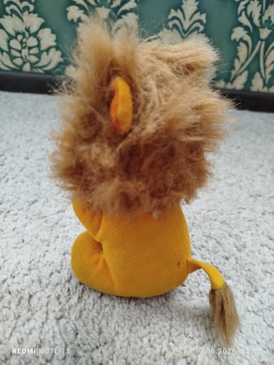 A close-up, high-angle shot shows the rear view of a plush toy lion sitting on a textured, light-colored carpet. The lion is primarily yellow with a shaggy, light brown mane. Its tail, also yellow with a brown tuft, extends to the right. The background features a patterned wallpaper with a dark teal base and cream-colored floral motifs, above a dark horizontal surface. The image is watermarked with "REDMI NOTE 13" and a date/time stamp "19.06.2026 10:53". The location in Асан-Чек, Kyrgyzstan is not depicted in the image itself, but is provided contextually.