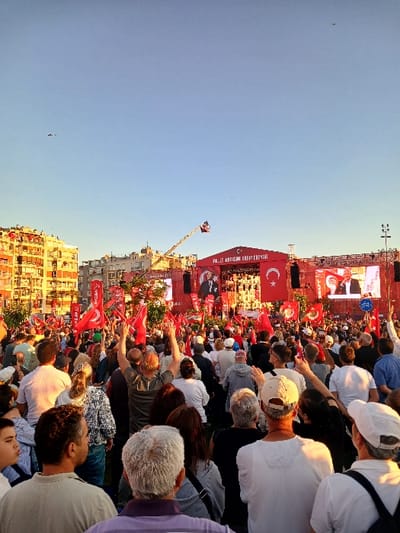The image shows a large crowd of people gathered in a public square in İzmir, Türkiye, at the coordinates 38.436, 27.141. The crowd is facing a large stage with a screen and several flags hanging above it. A man is giving a speech from the stage, facing the crowd. The people in the crowd are listening intently and waving Turkish flags. The square is surrounded by buildings. The sky is blue and clear, with a few birds flying overhead. The scene is bright and lively, with a festive atmosphere. Many people are holding red and white flags and signs, all facing the stage and the speaker.