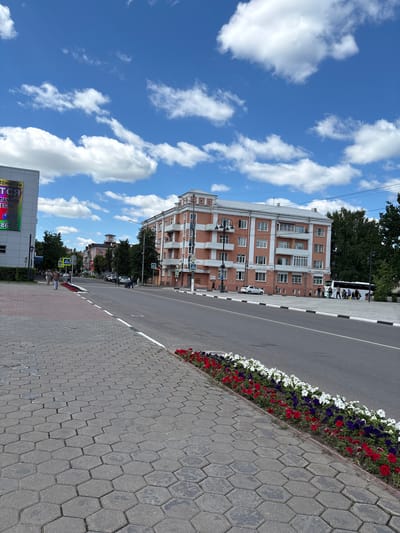 A wide, sunlit street in Noginsk, Russia, is depicted on a clear day with scattered white clouds. The street is bordered on the left by a hexagonal paver sidewalk and a modern, light-colored building with a large digital display. Further down the street, a pink, multi-story building with white trim and balconies dominates the right side of the frame. A white bus is parked in front of this building, and several people are visible near it, some appearing to be passengers with luggage. Further down the street, trees line the sidewalks, and various cars and pedestrians are present. A flower bed with red, white, and purple flowers runs along the edge of the sidewalk, separating it from the road. Traffic signs and pedestrian crossings are visible, indicating a well-trafficked urban area. The overall scene suggests a lively and functional city environment.