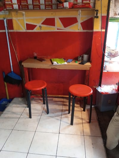 The image displays an interior scene in Ambakirano, Madagascar, featuring red walls with a horizontal yellow stripe and an irregular red and yellow geometric pattern above. A high wooden shelf is mounted on the wall, holding multiple rectangular boxes, predominantly red and white, labeled "DEPIUM". Below this, a narrow wooden counter is positioned against the red wall. On the counter, from left to right, are a clear plastic cup containing a dark liquid, a red smartphone, a crumpled green item resting on papers, a blue/purple smartphone, a small green globular object, a black smartphone, a white ceramic mug, and a bundle of dark sticks. Two red-topped stools with black metal legs are situated in front of the counter on a light-colored tiled floor with visible grout. To the far left, a blue and black broom with a white handle leans against the wall, alongside a yellow stick and a blue dustpan. The far right of the frame reveals a partial view of an opening, possibly a doorway or window, and a grey plastic bin with a white plastic bag liner. A person's lower leg, clad in light-colored trousers, is partially visible on the far right edge of the frame.