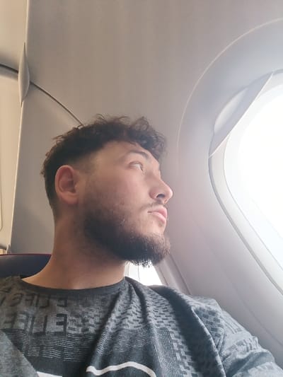 The scene depicts a male individual inside an aircraft cabin, contextually associated with the city of Saint-Louis, France. The subject, a male in his 20s or 30s with dark, curly hair featuring a faded cut on the sides, is shown in profile, looking upwards and to the right, gazing out an oval airplane window. He has a neatly trimmed beard and mustache. He is wearing a grey t-shirt with a subtle geometric pattern and white text, including the letters "E LIFE," visible on the chest area. The aircraft interior features light grey paneling and a portion of a dark-colored seat is visible to the left. The window shows a brightly lit, featureless exterior, consistent with an in-flight or on-ground view without discernible ground details. No other individuals or significant interactions are observed.