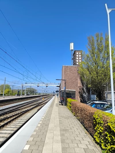 The image depicts a view of Uitgeest train station in the Netherlands. The scene is captured on a bright, clear day under a blue sky. On the left are several railway tracks with overhead power lines. A train is visible further down the tracks, approaching the station. The platform is paved with grey blocks and tactile paving. To the right of the platform is a low hedge with green and brown foliage. Behind the hedge are parked cars, including a blue hatchback. A brick building stands next to the cars, topped with a telecommunications antenna. A light pole stands on the right edge of the frame.