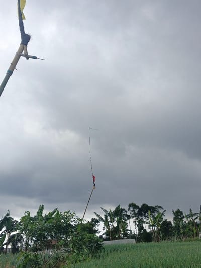 The image depicts an agricultural landscape in Ngọc Tụ, Vietnam, under a heavily overcast sky dominated by dark grey clouds. In the far left foreground, a section of a large bamboo pole extends vertically out of frame, featuring a dark wrapped material and a small, faded yellow fabric flag attached near its visible top. Below this flag, a white PVC pipe fitting with an attached horizontal metal rod protrudes from the pole. Centrally positioned in the midground, a second, slender pole, likely bamboo, stands upright, displaying white horizontal stripes and a small red piece of fabric tied approximately one-third up from its visible base. Near the top of this pole, a small blue rectangular object is affixed horizontally. The lower half of the frame reveals a cultivated field of dense, vibrant green, vertical plants, consistent with a scallion or onion crop, spanning the right side. To the left and center, various tall trees, including prominent banana trees with broad green leaves, are visible. A low-lying, elongated white structure, resembling a greenhouse or crop cover, is situated in the background behind the cultivated field. No human figures are present.