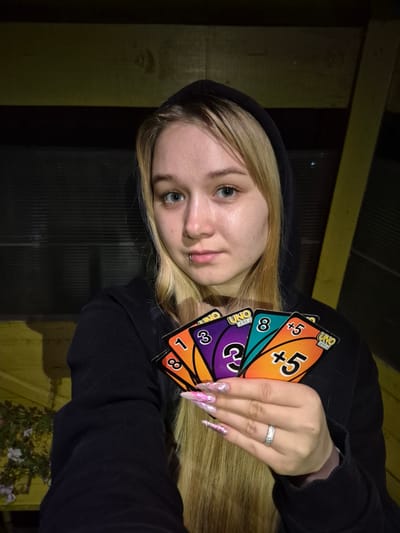 This media file is a photograph of a young woman, likely a teenager or young adult, holding several UNO cards. She is wearing a black hooded sweatshirt. Her blonde hair is long and falls over her shoulder. Her left hand, which is visible, is adorned with long, pointed, pink and iridescent acrylic nails, and she is wearing a ring on her left ring finger. The UNO cards are fanned out and held in her right hand. Visible cards include a "+5" card, an "8" card, a purple card with a "3", an orange card with a "1", and a card with a "2" on a blue background. The background appears to be outdoors, possibly on a porch or deck, with wooden beams and dark corrugated material visible. There are also some plants with flowers in the lower right corner of the frame. The lighting suggests it is evening or nighttime. The woman's expression is neutral. The location context is Sars, Russia.